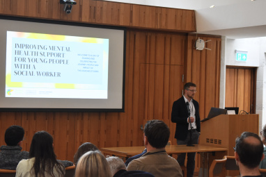 Man standing in front of screen addressing audience. Screen displays title of event: Improving mental health support for young people with a social worker