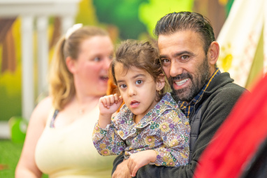 A father smiles while holding his daughter with a woman smiling in the background