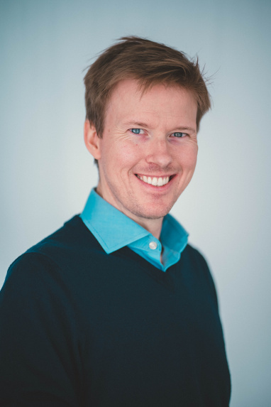 Man with short hair and light blue shirt and dark jumper smiling
