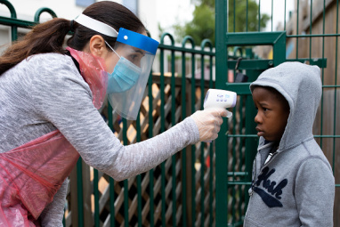 A child has their temperature checked by a teacher wearing protective clothing in a school playground