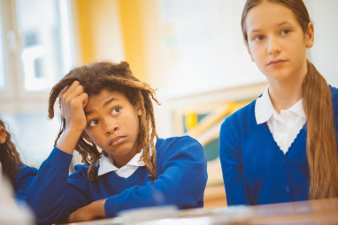 A Black schoolchild holds her head in her hand while sat next to a white classmate