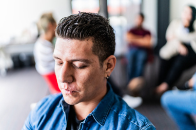 Man looking down with eyes closed with other adults talking in the background