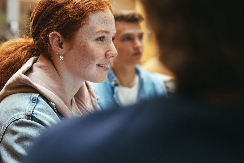 Profile of smiling teenage girl in a group wearing denim and butterfly earring with the silhouette of someone in foreground