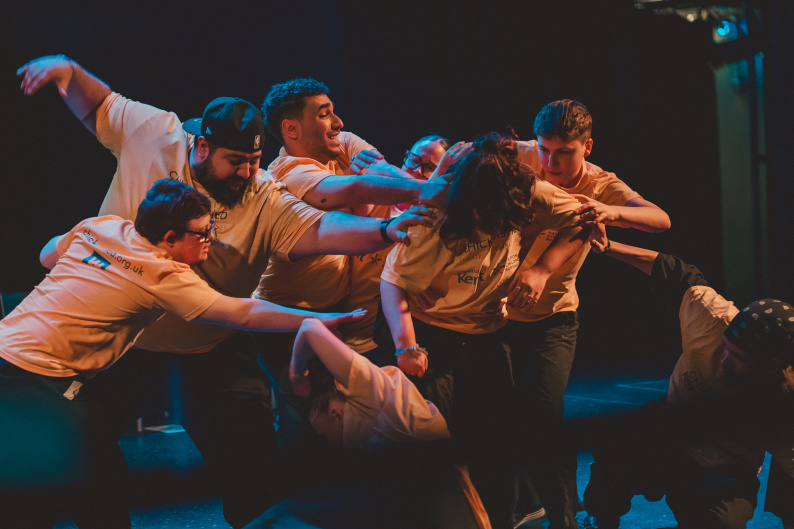 Young people acting in the final show surround a central performer, extending their arms towards her as her head is bowed down.