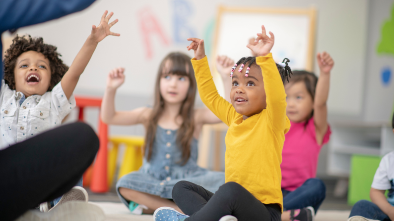 young children sat on carpet with their hands raised