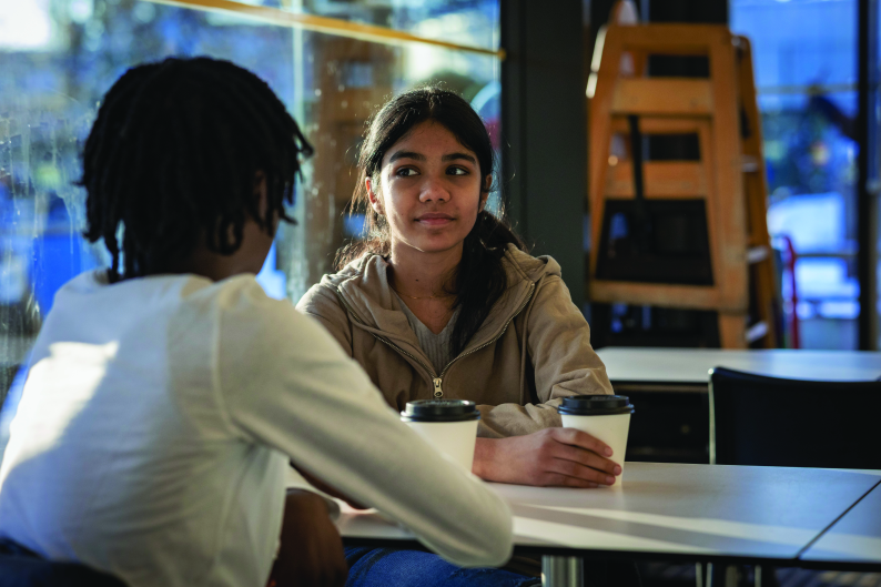 Two teenagers sitting at a table across each other, holding disposable coffee cups