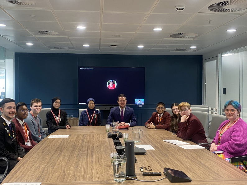Image of young people sat around a table looking at the camera and smiling, the Minister is sat in the middle of them.