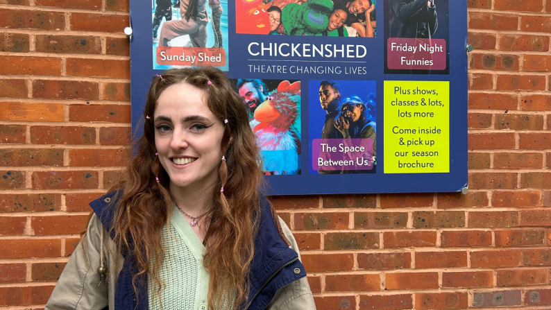 Hannah smiles as she stands outside the Theatre, of which we can see a notice board with the theatre's name and leaflets