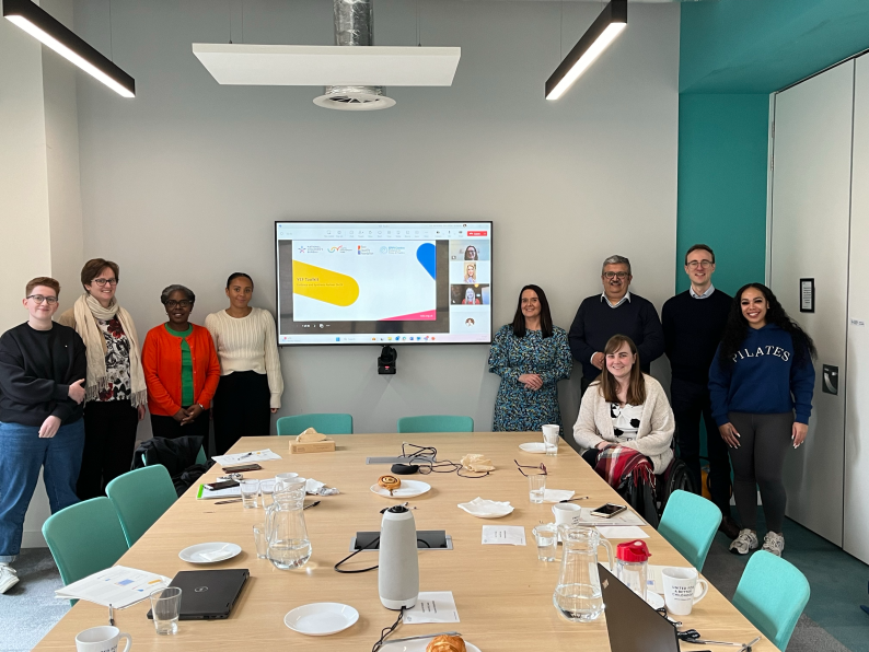 The team working on the toolkit pictured at the kick off meeting, nine people are in the room standing eitehr side of the tv screen where some of the team have joined in person.