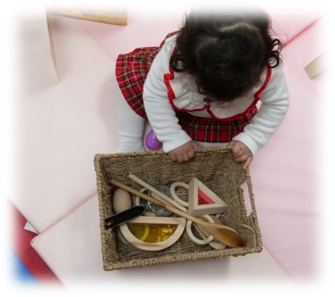 Image taken from above looking down at a small child looking in a box of wooden materials such as a wooden spoon and different shaped objects